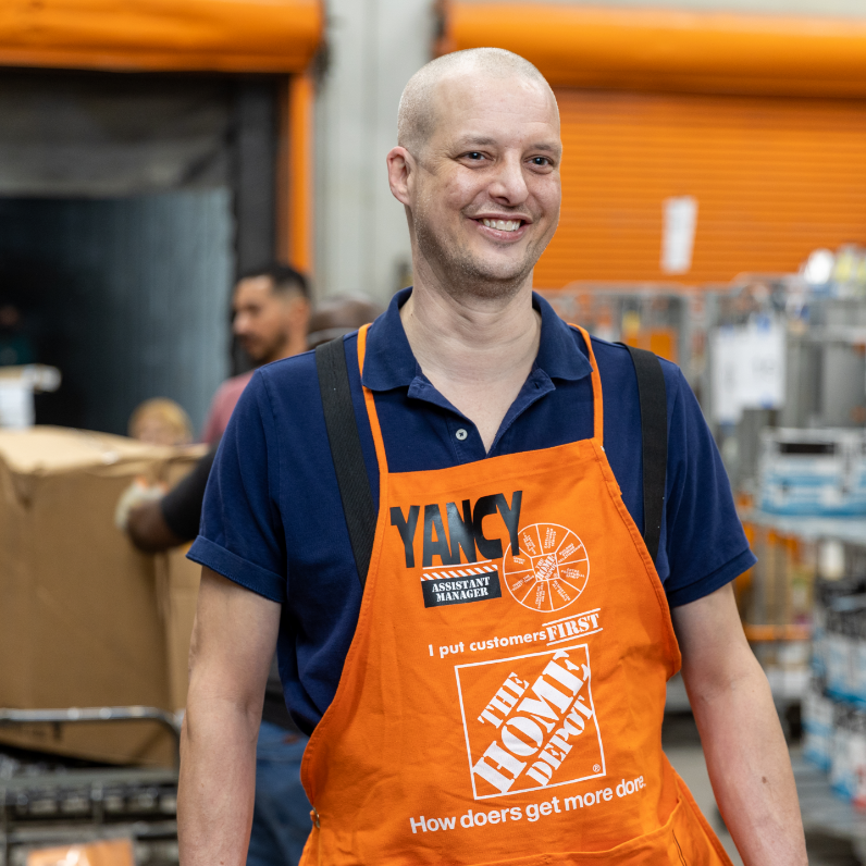 A middle-aged bald white man in an orange apron smiles while standing in a storage facility.